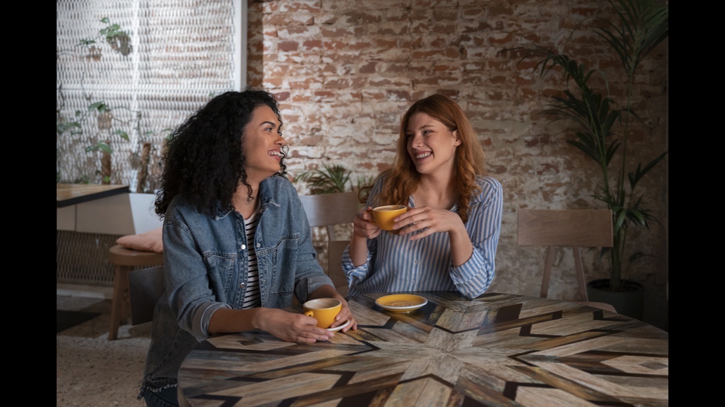 Dos personas riendo en una mesa de madera con tazas ocre, pared de ladrillo al fondo