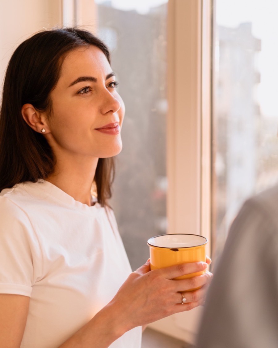 Mujer junto a una ventana sosteniendo una taza ocre mientras escucha a la persona frente a ella