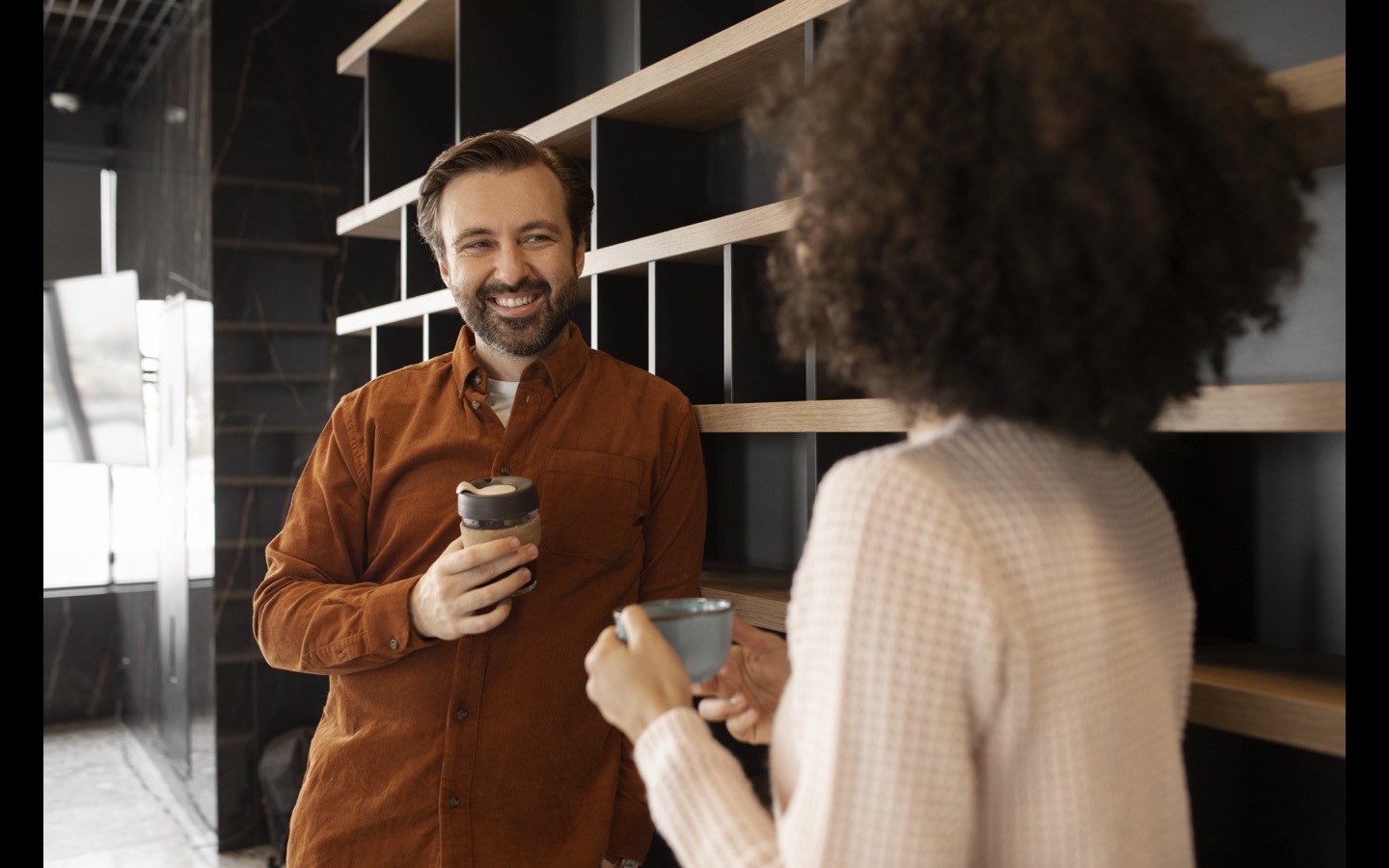 Dos personas conversando de pie con una taza en la mano, luz cálida de interior